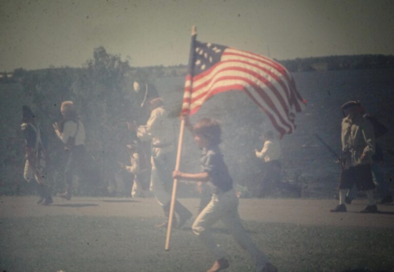 Teenage boy in period clothing runs across a grassy battlefield carrying a large American flag during a War of 1812 reenactment at Sackets Harbor, New York.