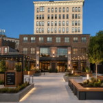 Concept rendering of a redeveloped JB Wise site in downtown Watertown, New York, showing a modern plaza with landscaped walkways, seating areas, storefronts, and renovated brick architecture