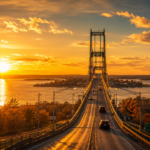 Wide-angle view of a large suspension bridge spanning the St. Lawrence River at sunrise, bathed in golden light. Vehicles travel across the bridge as the sun reflects off the water and illuminates the surrounding Thousand Islands landscape.