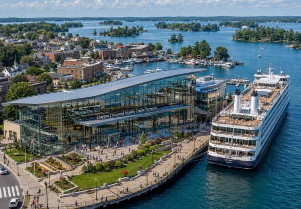 Concept image of a modern cruise terminal in Clayton, New York, featuring a large glass-fronted waterfront building, landscaped public promenade, docked cruise ship, marina, and Thousand Islands scenery in the background.