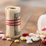 Prescription pills spilling from a bottle beside a rolled stack of U.S. currency on a wooden table.