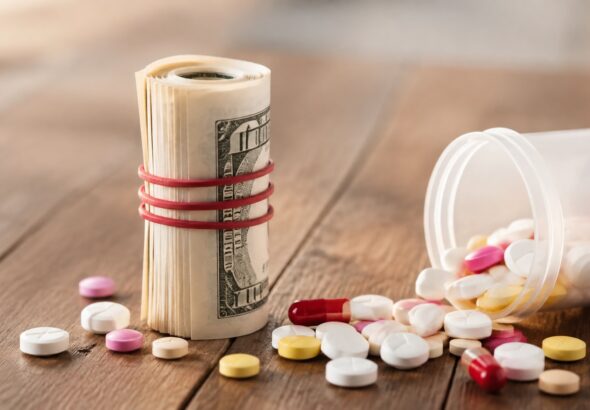 Prescription pills spilling from a bottle beside a rolled stack of U.S. currency on a wooden table.