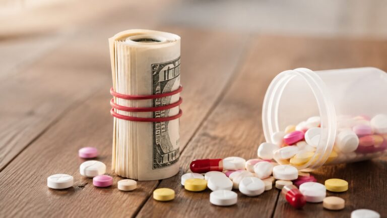 Prescription pills spilling from a bottle beside a rolled stack of U.S. currency on a wooden table.