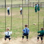 Amish children in traditional clothing playing on wooden swings in a rural schoolyard.