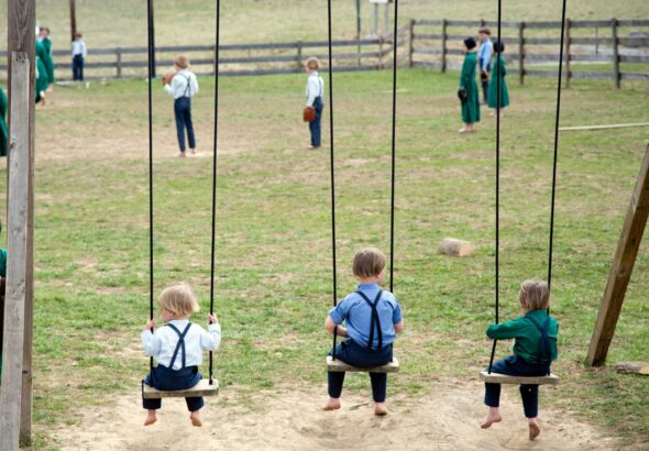 Amish children in traditional clothing playing on wooden swings in a rural schoolyard.