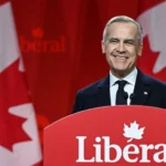 Canadian Prime Minister Mark Carney smiles behind a Liberal Party podium with large Canadian flags in the background.