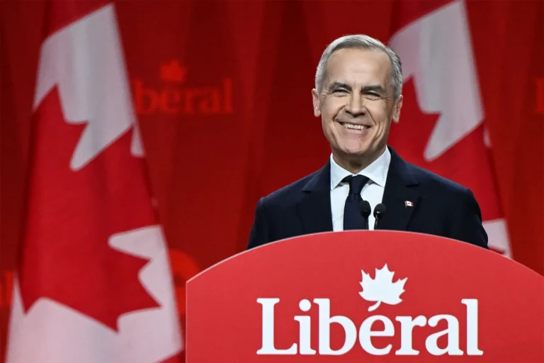 Canadian Prime Minister Mark Carney smiles behind a Liberal Party podium with large Canadian flags in the background.