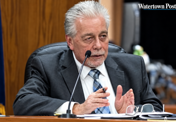 A suited city official speaks into a microphone while seated at a council desk with papers and glasses in front of him. A Watertown Post logo appears in the upper-right corner.