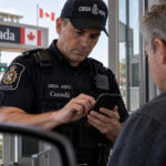 Canadian border officer examines an American traveler’s cellphone at a Canada border crossing as vehicles wait in the inspection lanes.